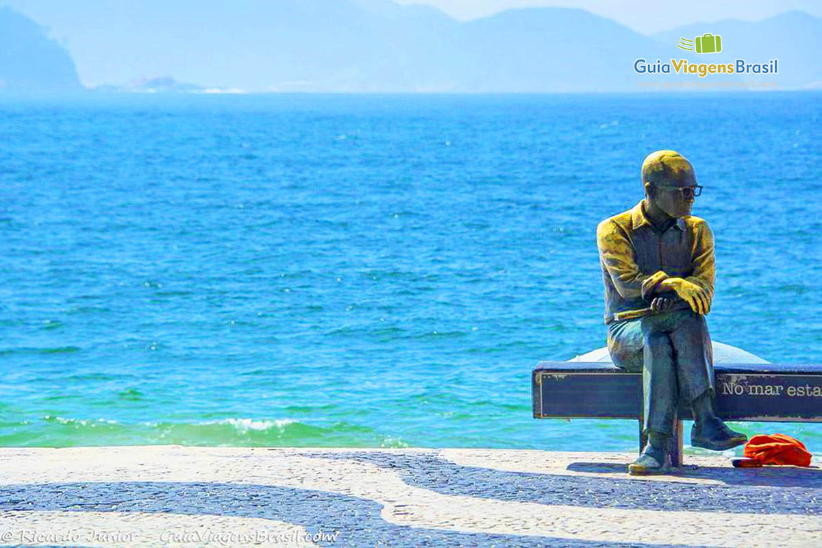 Imagem de Carlos Drumond de Andrade sentado em um banco na orla da Praia de Copacabana.